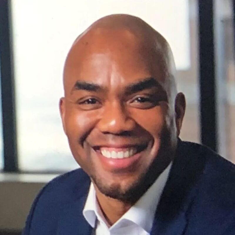 The image shows a smiling Black man with a shaved head. He is wearing a dark blue suit jacket and a white shirt. The background is blurred, but it appears to be an office setting with a window. The man has a warm and friendly expression.